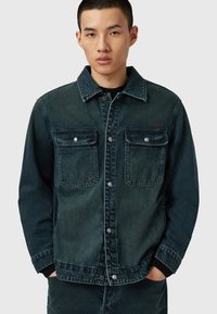 Tummasta farkkutakista, jossa on kaulus, napitussulku, kaksi rintataskua ja haalistunut pinta. Hihojen väri tummenee kohti hihansuita.