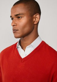 Muž v červeném svetr s výstřihem do V přes bílou kostkovanou košili s knoflíky, dívající se na stranu na pozadí s jednobarevným světlým pozadím.