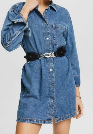 Frau trägt ein blaues Jeans-Knopfleid mit einem schwarzen Gürtel, der am Taillenbereich große silberne Kettenglieder aufweist.