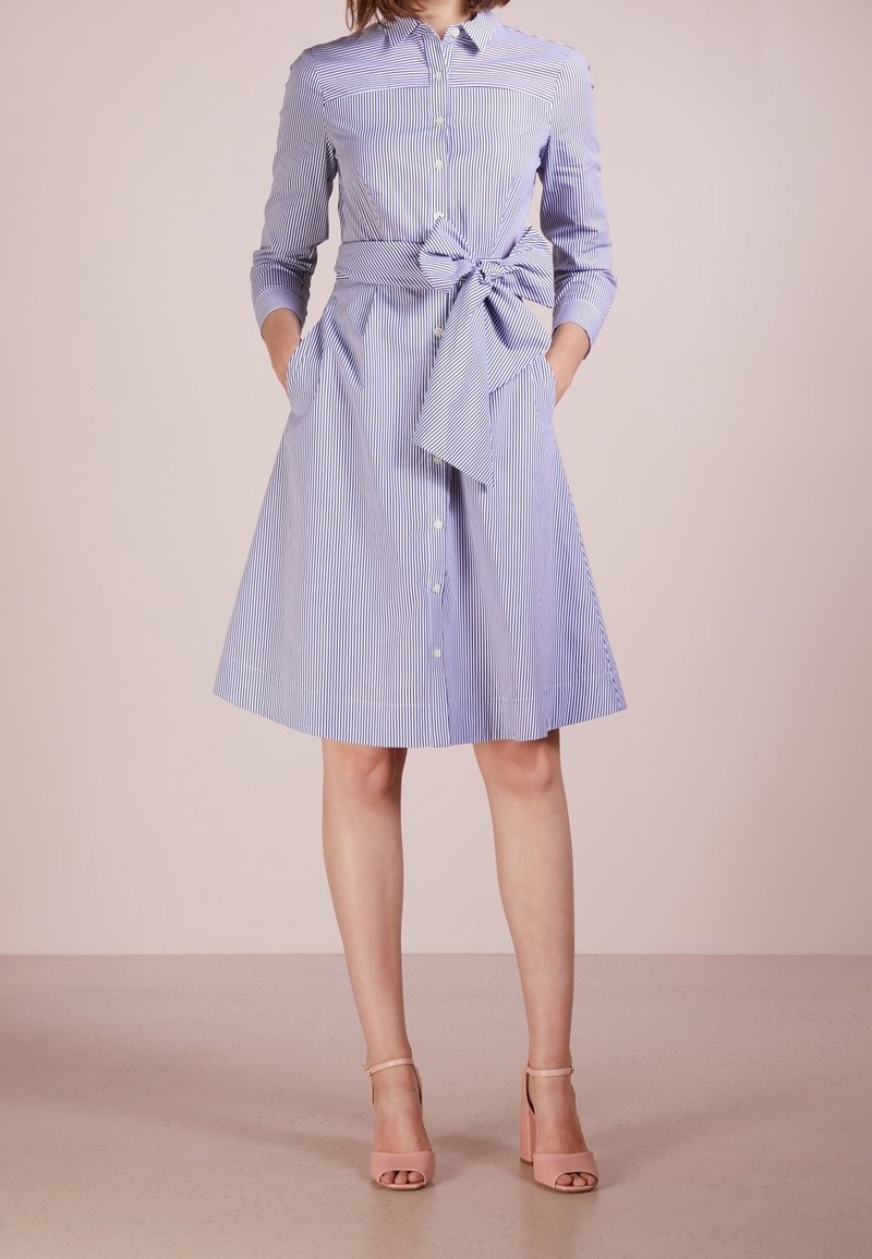 Femme portant une robe chemise rayée bleu et blanc jusqu'aux genoux avec un grand nœud à la taille et des talons nude à bride autour de la cheville.