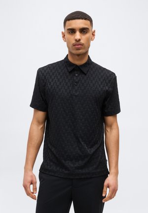 Młody mężczyzna ubrany w czarną koszulkę polo z krótkim rękawem o subtelnym wzorze w kratę, stojący na tle jednolitego jasnego tła.