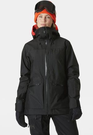 Schwarze isolierte Skijacke mit hohem Kragen, Reißverschluss und Taschen. Aus wasserdichtem Material und mit verstellbarem Kapuze. Kombiniert mit Handschuhen.