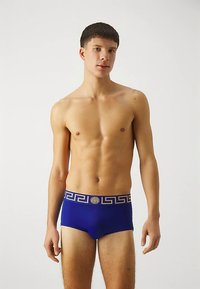 Blue swim trunks feature a gold Greek key pattern on the waistband and a central branded accent. Made of smooth, stretchy material.