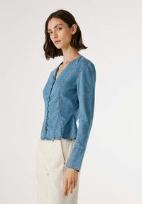 Gaišas zila džinsu blūze ar garām piedurknēm, V veida kaklu, priekšējo pogu aizdari un pieguļošu siluetu. Iezīmē redzamas šuves un īsāku apakšu.