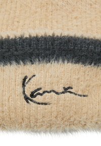 Bézs kötött anyag bolyhos textúrával, vízszintes sötétszürke csíkkal és fekete hímzett mintával az alján.