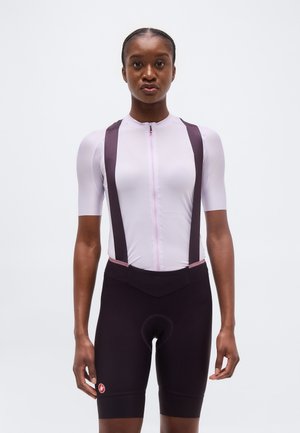 Sieviete stāv, skatoties uz priekšu, tērpusies gaiši rozā īsroku velosipēda krekliņā un melnos biksēs ar lencēm, baltā vienkāršā fonā.