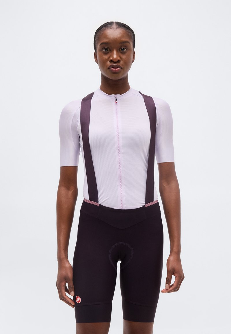 Γυναίκα που στέκεται όρθια προς τα εμπρός φορώντας κοντομάνικη ποδηλατική φανέλα ανοιχτού ροζ χρώματος με μαύρο σορτς bib, σε απλό λευκό φόντο.