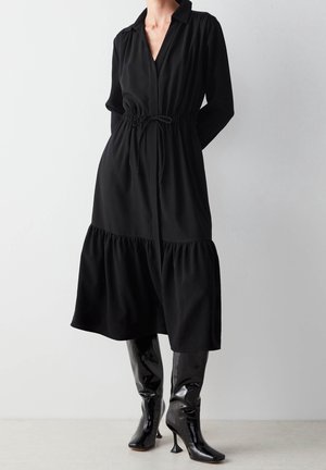 Femme portant une robe midi noire à manches longues avec une taille élastique et un ourlet à volants, associée à des bottes noires brillantes à talons montant jusqu'aux genoux.