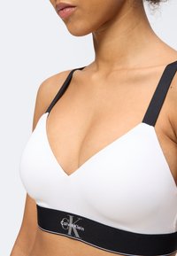 Γυναίκα που φοράει λευκό αθλητικό σουτιέν Calvin Klein με μαύρους ιμάντες και φαρδιά μαύρη ταινία κάτω με λογότυπο, εμφανιζόμενη από τον λαιμό έως το μέσο της κοιλιάς.