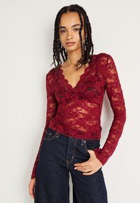 Blusa de renda burgundy com mangas longas, decote em V com bordas onduladas e padrão floral, combinada com jeans de ganga escura. Textura transparente.