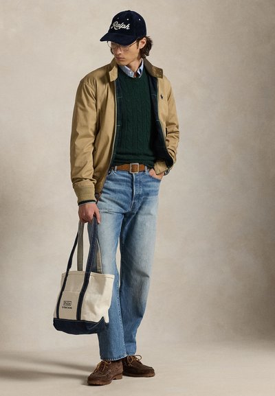 Noor mees, kandes beežikat jope, rohelist kampsunit, siniseid teksapükse ja tumesinist Ralphi nokamütsi, hoides vasakus käes beeži ja tumesinist kandekotti.