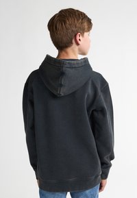 Zwarte hoodie met een donkerdere, licht vervaagde kap, met een relaxte pasvorm en geribbelde manchetten. De achtergrond is eenvoudig wit, wat het kledingstuk benadrukt.