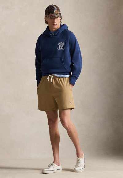 Μπλε ναυτικό φούτερ με λογότυπο, μπεζ σορτς με ελαστική μέση και λευκό κορδόνι, λευκά αθλητικά παπούτσια και καρό καπέλο. Ομαλές υφές, casual σχεδίαση.
