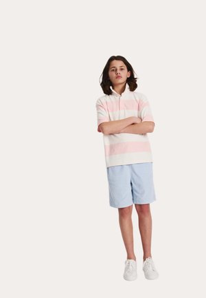 Persona joven de pie con los brazos cruzados, vistiendo una camisa polo de rayas rosas y blancas, pantalones cortos azules y zapatillas blancas, sobre un fondo liso.