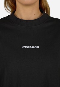 Schwarzes Baumwoll-T-Shirt mit klassischem Rundhalsausschnitt, ausgestattet mit weißem, gesticktem "PEGADOR"-Logo auf der Vorderseite. Glatte Textur, minimalistisches Design.