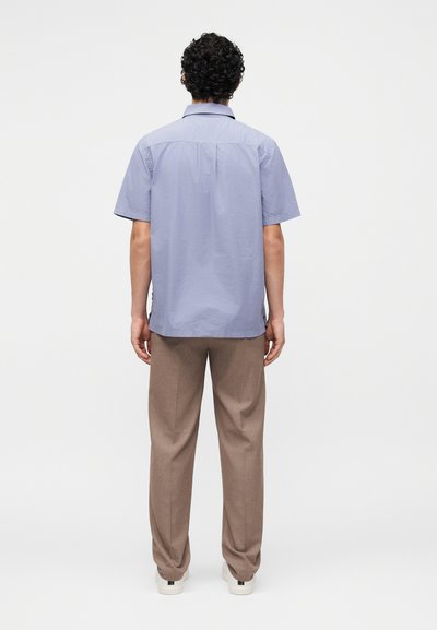 Άνδρας με σγουρά σκούρα μαλλιά στέκεται με το πρόσωπο προς τα πίσω, φοράει ανοιχτό μπλε κοντομάνικο πουκάμισο, καφέ παντελόνια και λευκά αθλητικά παπούτσια σε ένα απλό φόντο.