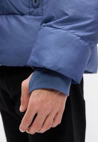 Manica di una giacca a vento blu con texture trapuntata, polsino elasticizzato in blu abbinato al polso, mano appoggiata su un tessuto nero.