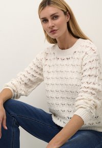 Maglione lavorato a maglia di colore crema, con un motivo aperto e testurizzato e colletto a coste. Vestibilità ampia con maniche larghe, abbinato a jeans blu scuri.