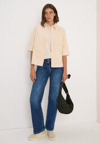 Ljusbeige oversized skjorta med korta ärmar och en bröstficka, kombinerad med blå utsvängda jeans och krämfärgade sneakers. Bär en mörkgrön väska.