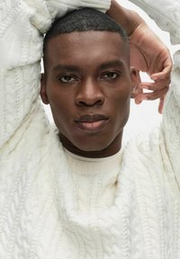 Lähikuva lyhyttukkaisesta miehestä, joka pukeutunut valkoiseen kaapelikuvioiseen neulepaitaan ja nostaa kädet päänsä yläpuolelle yksiväristä vaaleaa taustaa vasten.