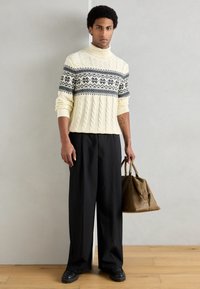 Suéter branco de malha com padrão preto; calças largas pretas; a segurar uma mala de couro castanha; em pé sobre um chão de madeira.