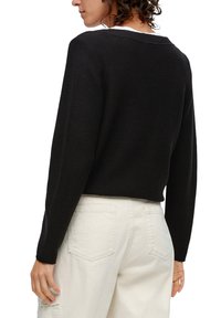 Schwarzer, gerippter Strickpullover mit einem V-förmigen Rückenausschnitt, langen Ärmeln und einem verkürzten Schnitt, kombiniert mit weißen, hochgeschnittenen Hosen.