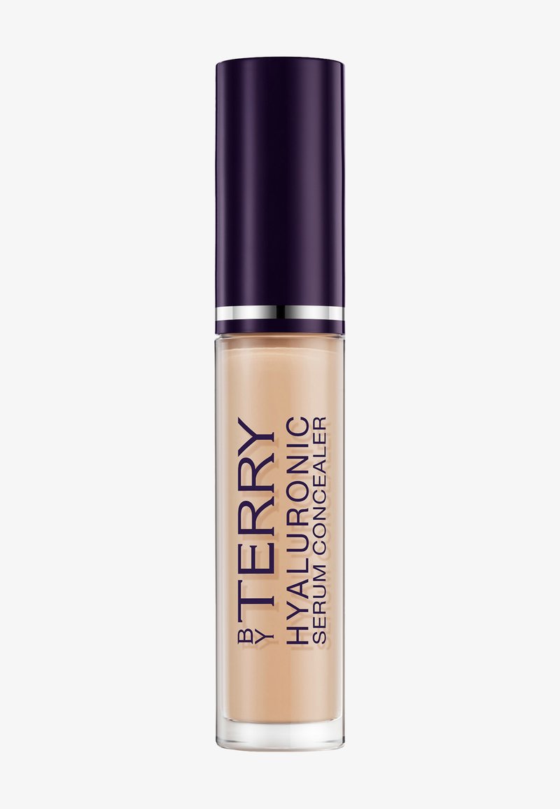 Gennemsigtig cylindrisk flaske med beige By Terry Hyaluronic Serum Concealer med mørkelilla låg og sølvbånd på hvid baggrund.