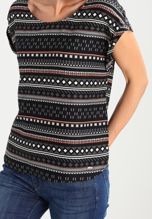 Kvinde iført en sort kortærmet bluse med hvide og røde geometriske mønstre parret med blå jeans, hånd i baglommen.