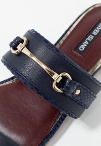 Sandale bleu marine à enfiler avec semelle intérieure grenat, détail mors doré et accents de brides texturées.