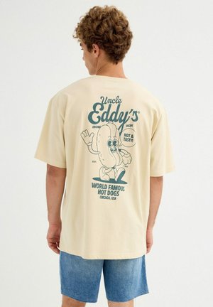 Joven con cabello rizado que lleva una camiseta beige con un dibujo animado de un perro caliente y el texto "Los Perritos Calientes Famosos en el Mundo de Tío Eddy" en la parte trasera.