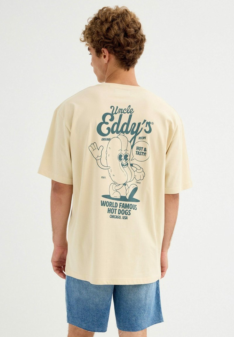 Joven con cabello rizado que lleva una camiseta beige con un dibujo animado de un perro caliente y el texto "Los Perritos Calientes Famosos en el Mundo de Tío Eddy" en la parte trasera.