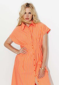 Robe à boutons orange rayée avec des manches courtes, dotée d'un col, de boutons sur le devant et d'une grande fente sur le côté. Texture de tissu lisse.