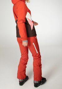 Personne portant un pantalon de ski rouge avec une poche zippée sur le côté, des chaussures de ski noires et une veste multicolore rouge, rose, blanche et noire, debout de côté.