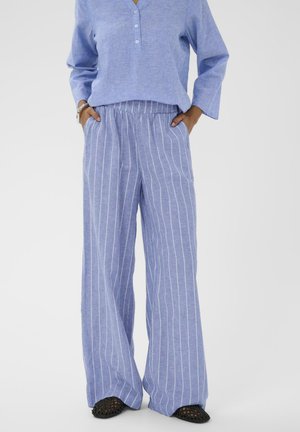 Femme portant un pantalon ample bleu clair à rayures blanches verticales et un haut à manches longues bleu clair assorti, les mains dans les poches.