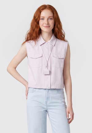 Junge Frau mit langen roten Haaren, die ein ärmelloses hellrosa Shirt und hellblaue Jeans trägt und mit einer Hand in der Hüfte steht.