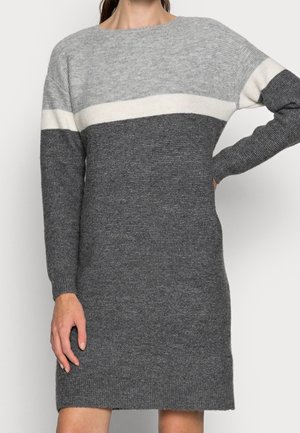 Długi sweterkowy sweter z długim rękawem w poziome paski w jasnoszarym, białym i ciemnoszarym kolorze, noszony przez stojącą osobę z ręką na biodrze.