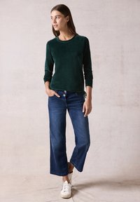 Haut en velours vert à manches longues avec poignets ajustés, associé à un jean en denim sombre à jambes larges et des baskets blanches décontractées.