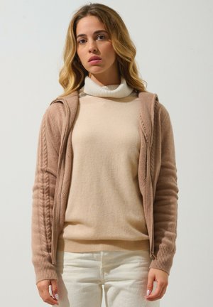 Beige gestrickter Pullover mit Rollkragen unter einer hellbraunen Strickjacke mit Zopfmuster und einem Reißverschluss. Weiße Hosen vervollständigen das Outfit.