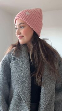 Căciulă roz din tricot cu dungi și finisaj texturat, cu o etichetă mică cusută, purtată cu o haină gri din fleece și un top negru. Părul este lung și ondulat.