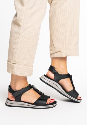 Feet in beige rolled-up pants wearing black open-toe sandals with thick straps and rubber soles, showing red-painted toenails.