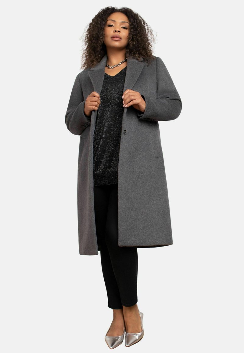 Cappotto in misto lana grigio con collo a scialle, chiusura a un bottone e tasche, abbinato a un top nero in paillettes e pantaloni neri, scarpe argentate.