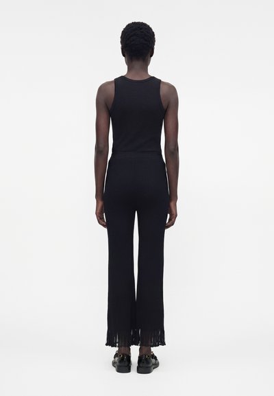 Οπίσθια όψη ενός όρθιου ατόμου που φοράει μαύρο αμάνικο τοπ, μαύρο πλισέ παντελόνι με φαρδιά πόδια και μαύρα παπούτσια με χρυσές λεπτομέρειες.