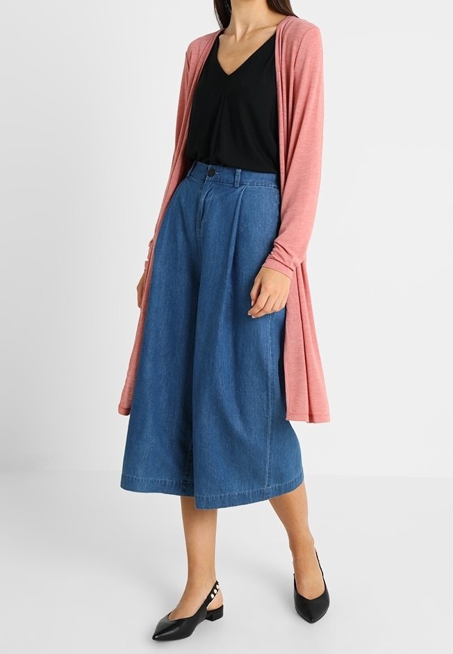 Cardigan rose léger à manches longues porté sur un haut noir, associé à un pantalon culotte en denim bleu à jambes larges et des ballerines noires avec des détails en boucle.