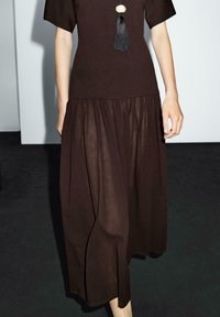 Braunes, knielanges Kleid aus strukturiertem Stoff; verfügt über kurze Ärmel, einen gerafften Rock und einen dekorativen Quastenakzent am Ausschnitt.