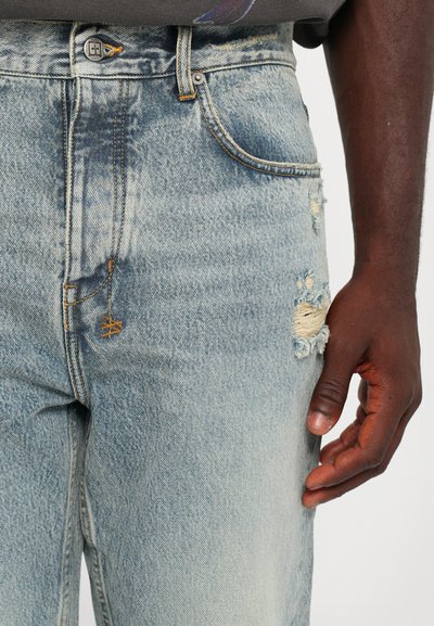 Jeans din denim de culoare albastru deschis cu un finisaj uzat. Prezintă închidere cu buton, cusături bej și mai multe buzunare. Distincție prin desfăcuturi și găuri vizibile.