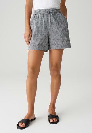 Kvinde iført grå og hvide stribede shorts med snøre, hvid top og sorte slip-on sandaler, stående mod en ensfarvet lys baggrund.