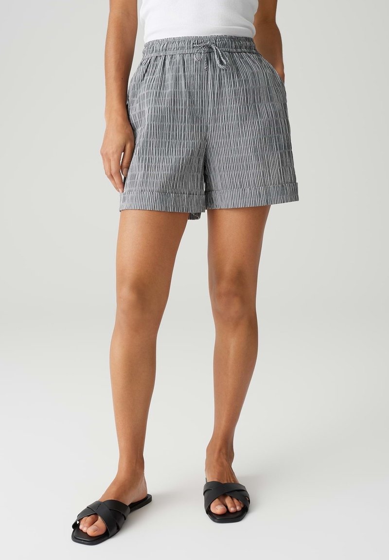 Femme portant un short rayé gris et blanc à cordon, un haut blanc et des sandales noires slip-on, debout devant un fond clair uni.