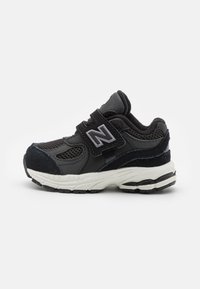 Chaussure de sport noire avec une tige en mesh et en daim. Dotée d'une sangle Velcro, d'une semelle blanche épaisse et de accents gris sur le logo et les détails.