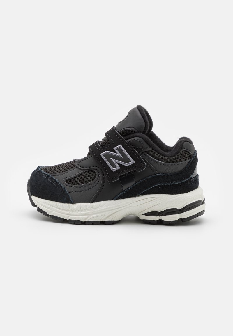 Chaussure de sport noire avec une tige en mesh et en daim. Dotée d'une sangle Velcro, d'une semelle blanche épaisse et de accents gris sur le logo et les détails.