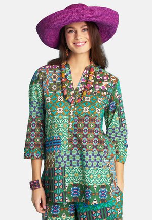 Smiling woman wearing a large purple hat, colorful patterned top and pants, beaded necklace, and matching bracelet, standing with hand in pocket.
